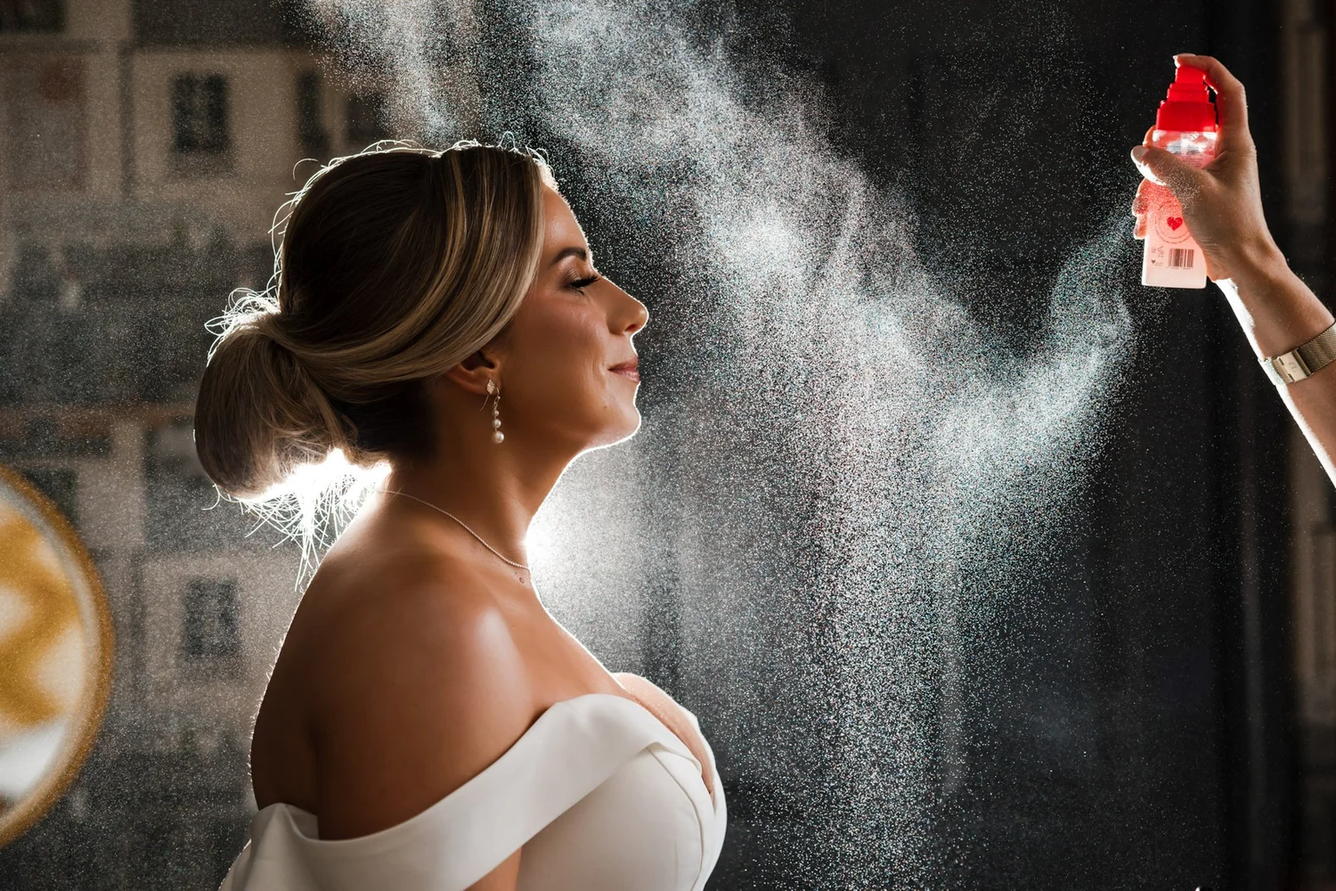A bride in profile with her hair pinned up, backlit hairspray drifting across a dark window behind her