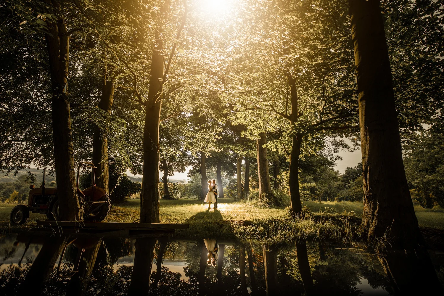 A bride and groom standing in low golden sunlight among tall trees, reflected in still water in the foreground, an old tractor to one side
